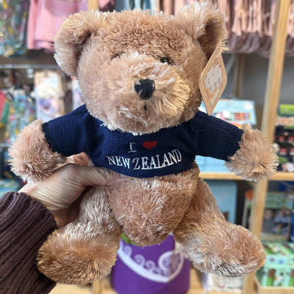 Teddy bear wearing a 'I ❤️ New Zealand' sweater held by a person in a store setting.